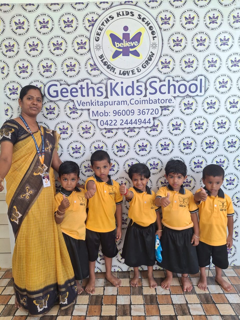 A teacher with little students in front of the Geeths Kids School logo wall
